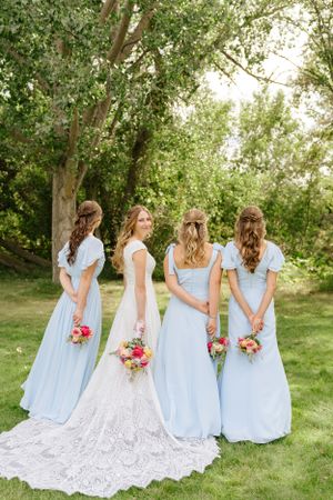 Bridal party in light blue dresses and white gown pose with bouquets facing away in garden setting.