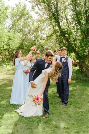 Wedding party celebrates with a joyful dip pose on a lush green lawn surrounded by trees.