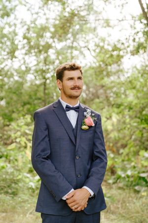 Groom in navy suit with bow tie and boutonniere poses in garden setting.