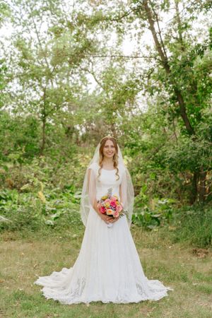 Bride in lace wedding gown holds colorful bouquet while standing in natural outdoor setting.