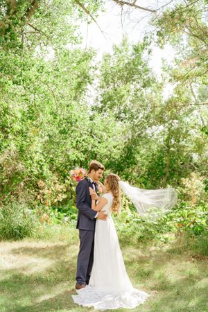 A romantic wedding photo of a couple embracing outdoors under lush green trees with a flowing veil at Taber MD.