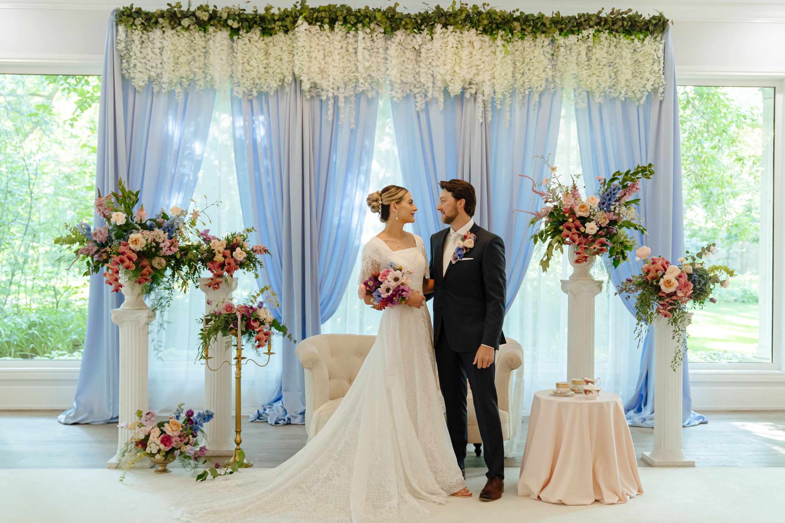 Wedding ceremony setup with light blue draping and floral arch featuring pink and white flowers.