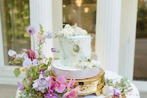 Elegant white wedding cake decorated with pink and purple flowers on gold stand.