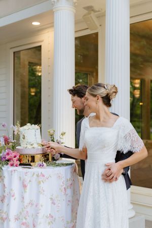 Couple cutting elegant wedding cake decorated with pink flowers on floral tablecloth by white columns.