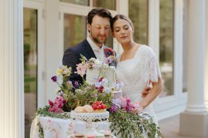 Couple standing by ornate wedding cake and floral arrangements on draped table with white columns.