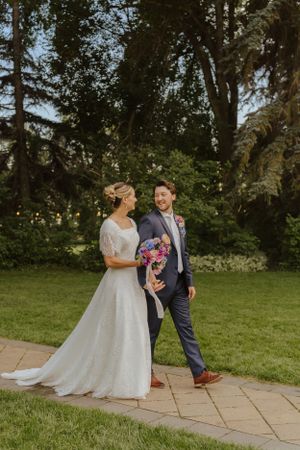 Wedding couple poses on garden path with colorful bouquet against green tree backdrop.