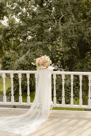 A long white bridal veil with peach flowers drapes elegantly across a white wooden railing in a garden setting.
