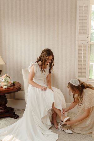Wedding dress preparation scene with shoes and accessories being fitted in a sunlit room.