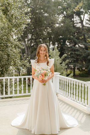 A white wedding dress flows elegantly on a sunny porch with white railings and green trees in the background.