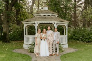Wedding party poses together in a white gazebo surrounded by lush green trees.