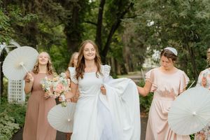 Wedding party in pastel dresses holding white parasols in a garden setting.