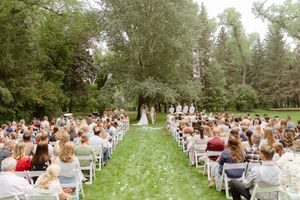 Outdoor wedding ceremony with rows of guests seated on white chairs along a tree-lined path.