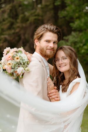 A couple wrapped in flowing white fabric share tender moments against a backdrop of green foliage.