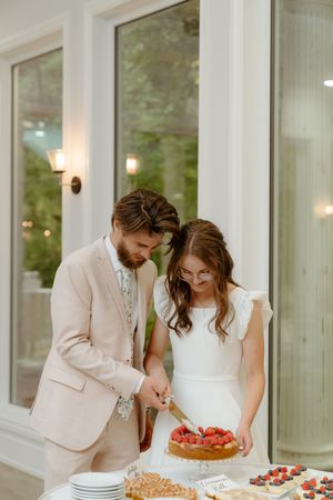 A couple cuts their wedding cake together at an elegant indoor reception venue with white decor.