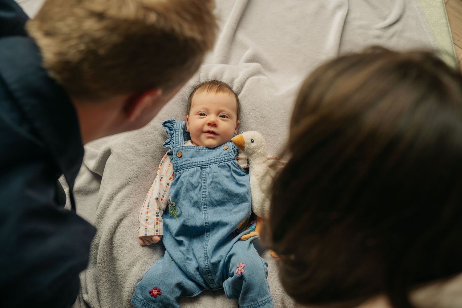 A baby wearing blue denim overalls smiles while sitting on a couch between two people.