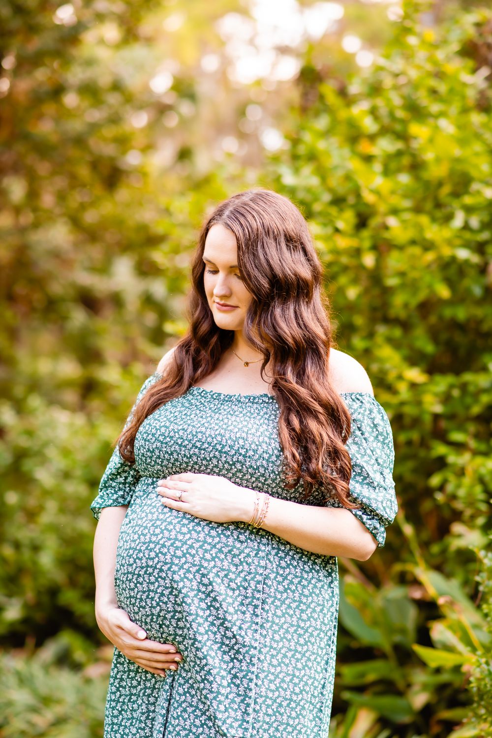Expectant mother in teal floral dress poses outdoors during maternity photoshoot in lush natural setting.
