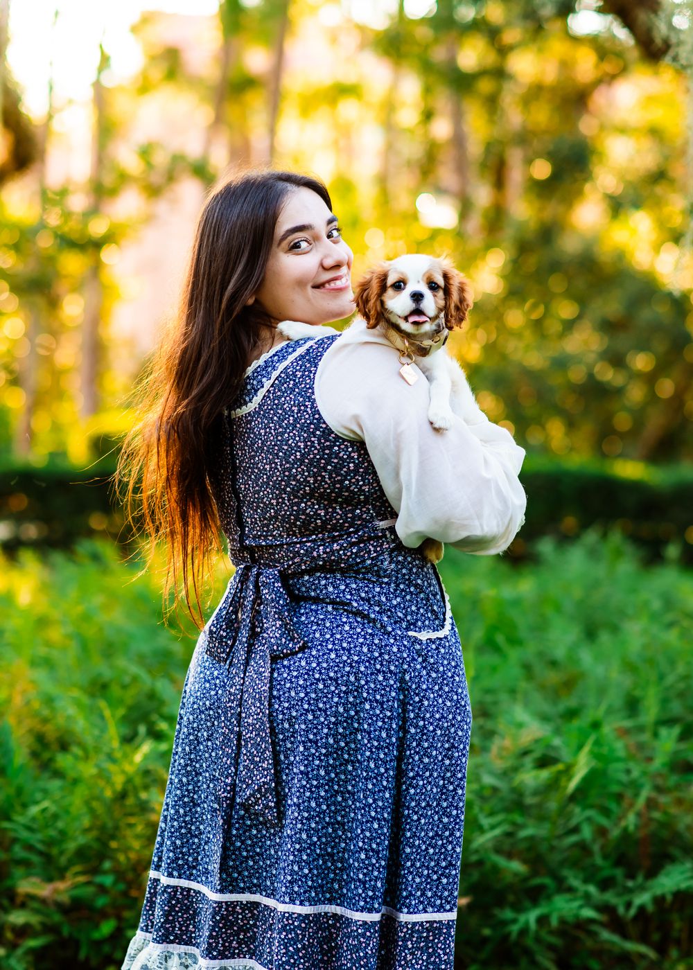 A young woman in a blue floral dress holds a Cavalier King Charles Spaniel in a white bag outdoors.