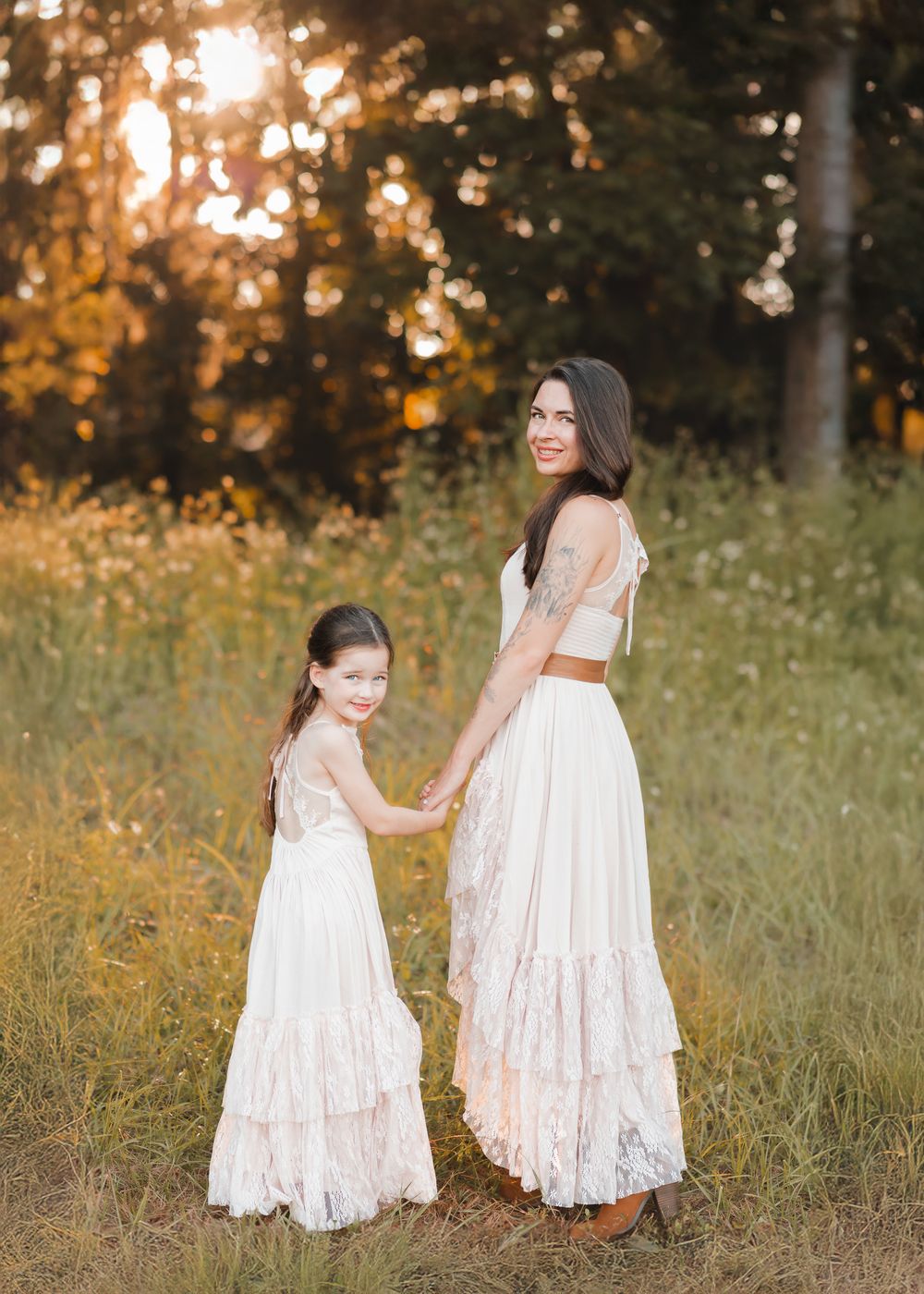 Two children in white lace dresses walk together in a sunlit meadow during golden hour.