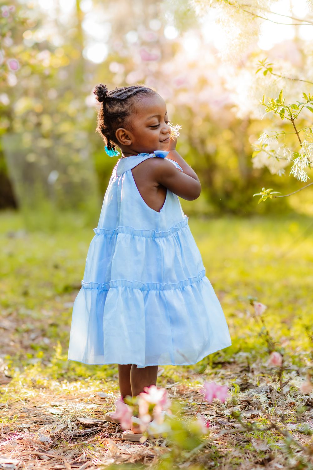 Young Black girl in blue dress smelling spring flowers outdoors in sunlit garden.