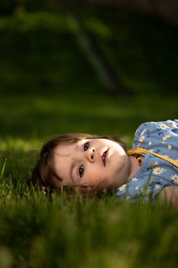 A young child lying on lush green grass, wearing a blue floral outfit, gazing upward in a serene outdoor setting.
