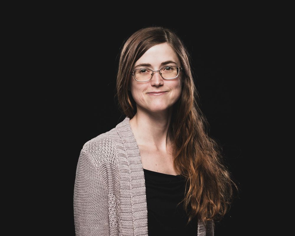 Young woman with long brown hair and glasses wearing a gray cardigan, smiling against a dark background.