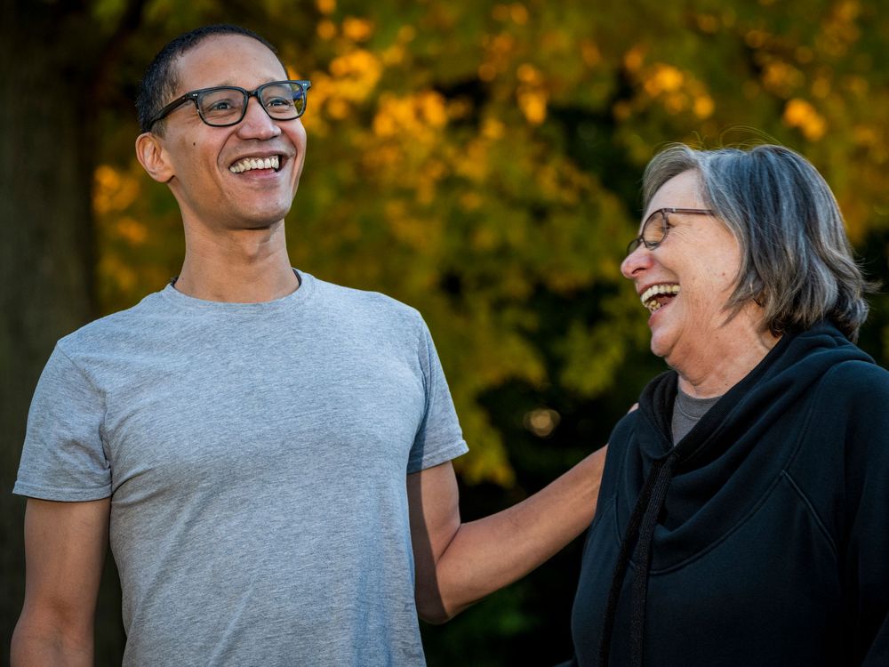 Two people laughing together outdoors in autumn, man in gray t-shirt and older woman in black jacket sharing a joyful moment.