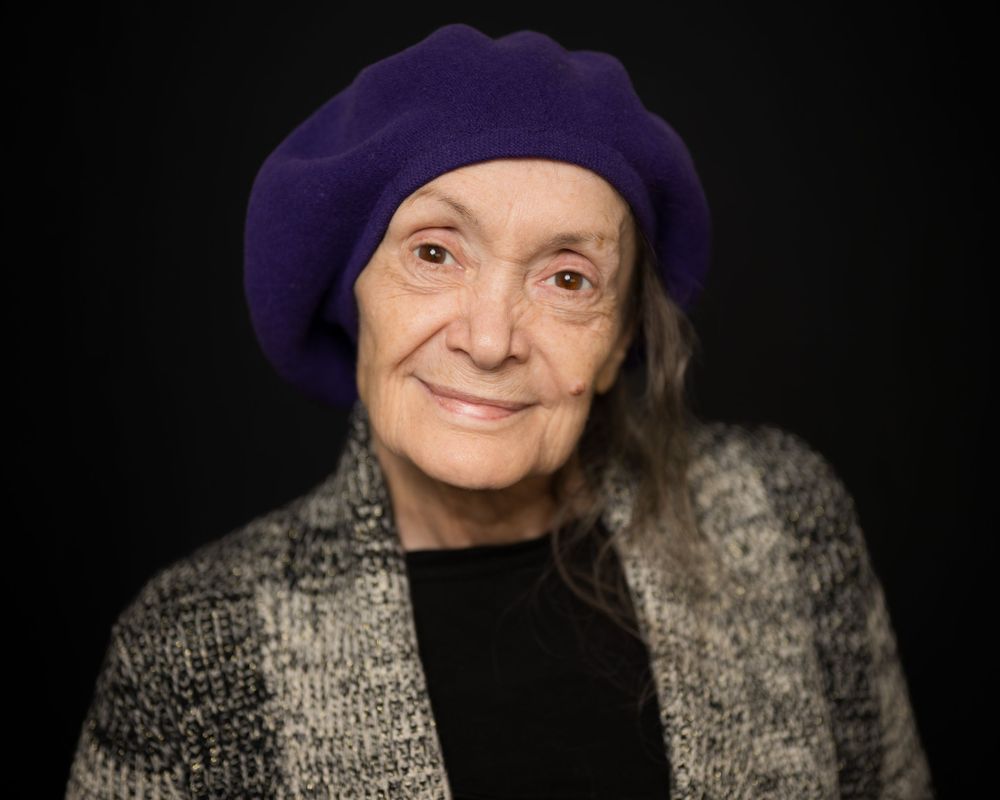 Elderly woman wearing a purple beret and checkered jacket, posing against a dark background.
