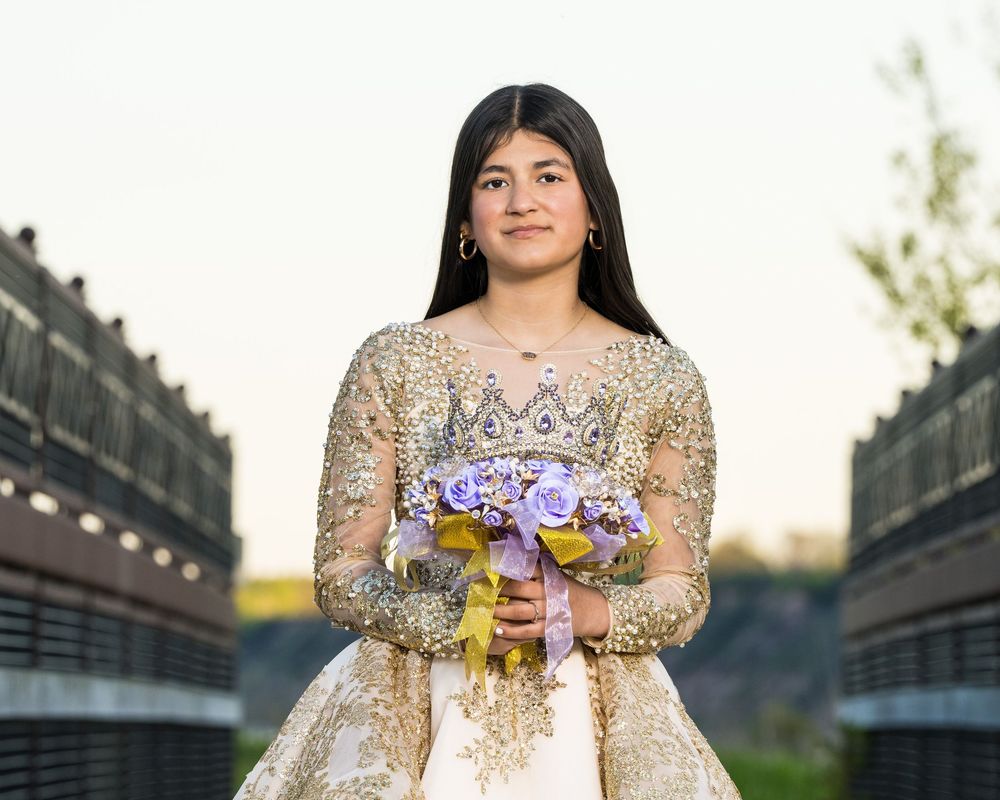 Young woman in gold quinceañera gown holding purple floral bouquet, posing outdoors at golden hour.
