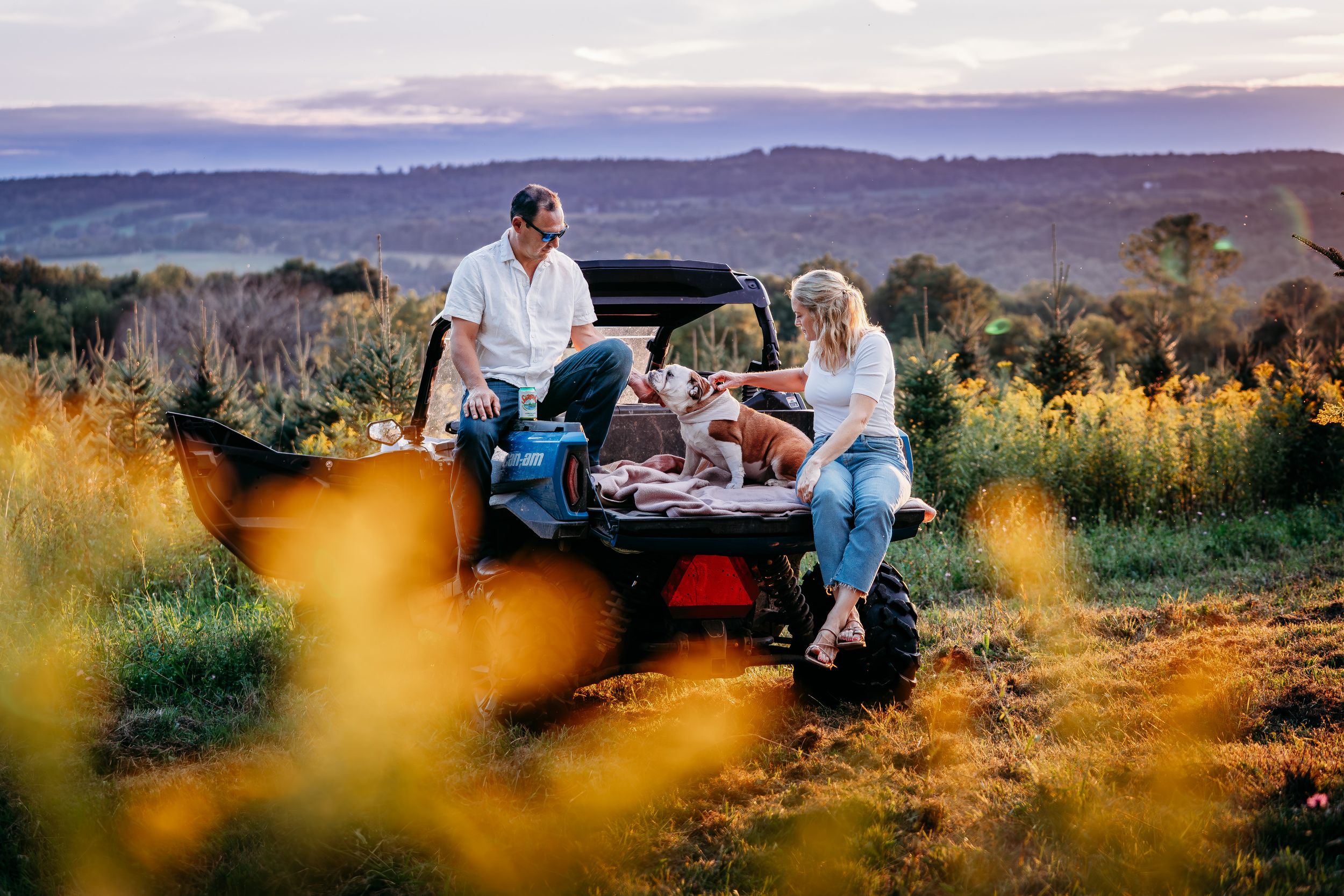 Couple relaxing by a UTV at golden hour in a scenic field with rolling hills and wildflowers in the background.