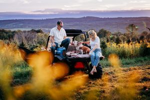Couple relaxing by a UTV at golden hour in a scenic field with rolling hills and wildflowers in the background.