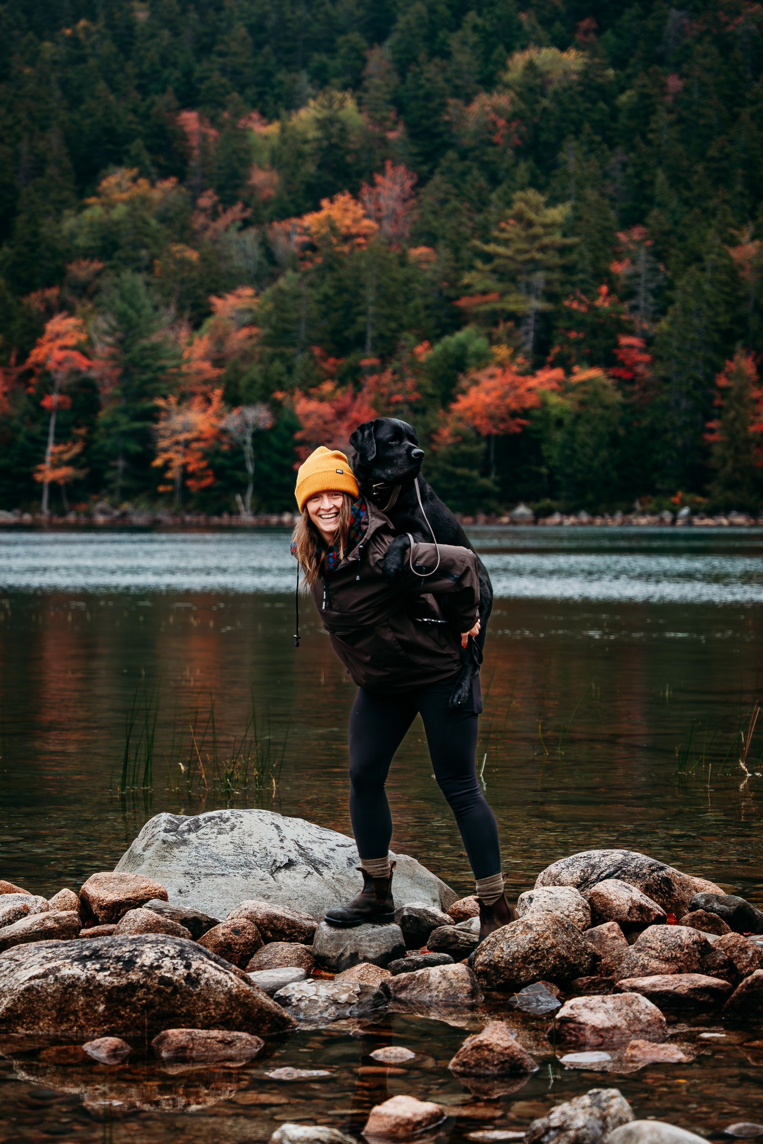 Woman in an orange beanie carries a black dog on her back while standing on lakeside rocks, with colorful autumn foliage behind.