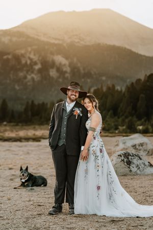 A couple poses with their dog against a mountain backdrop during a wedding photoshoot at sunset.