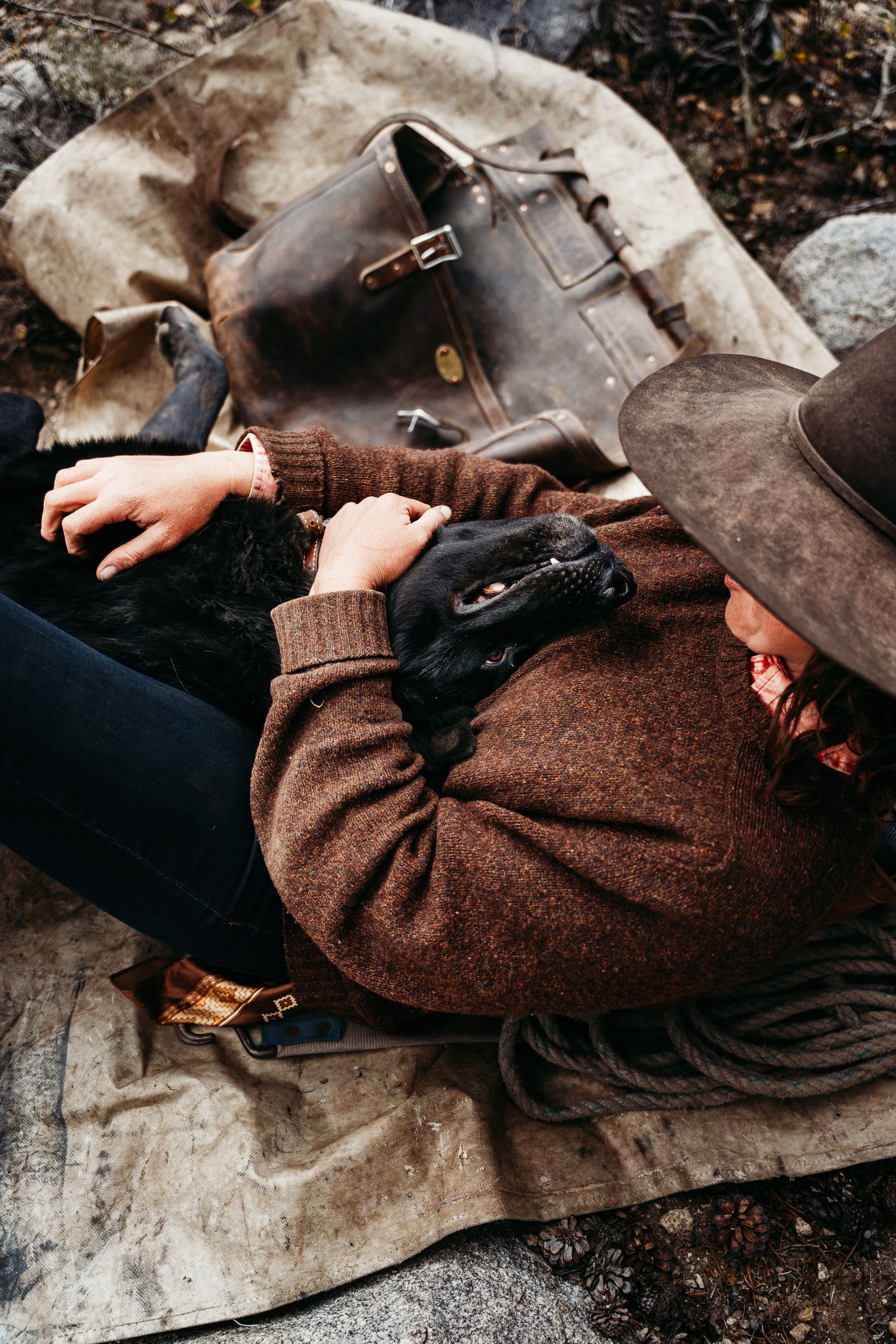 Person in a brown wool sweater and felt hat rests on a canvas tarp, cuddling a black dog beside a leather bag and coiled rope.