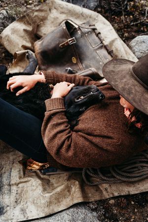 Person in a brown wool sweater and felt hat rests on a canvas tarp, cuddling a black dog beside a leather bag and coiled rope.