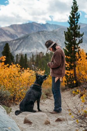 Person in a wide-brim hat and brown sweater stands on a mountain trail beside a black dog, surrounded by golden autumn foliage.