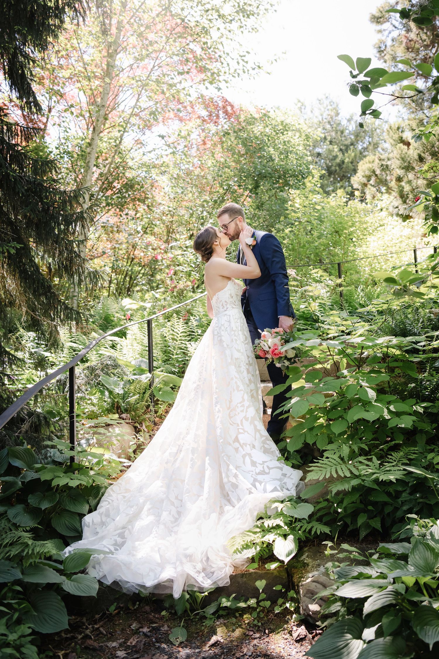 A couple embraces on garden steps surrounded by lush greenery, with the bride wearing a flowing white lace gown with long train.