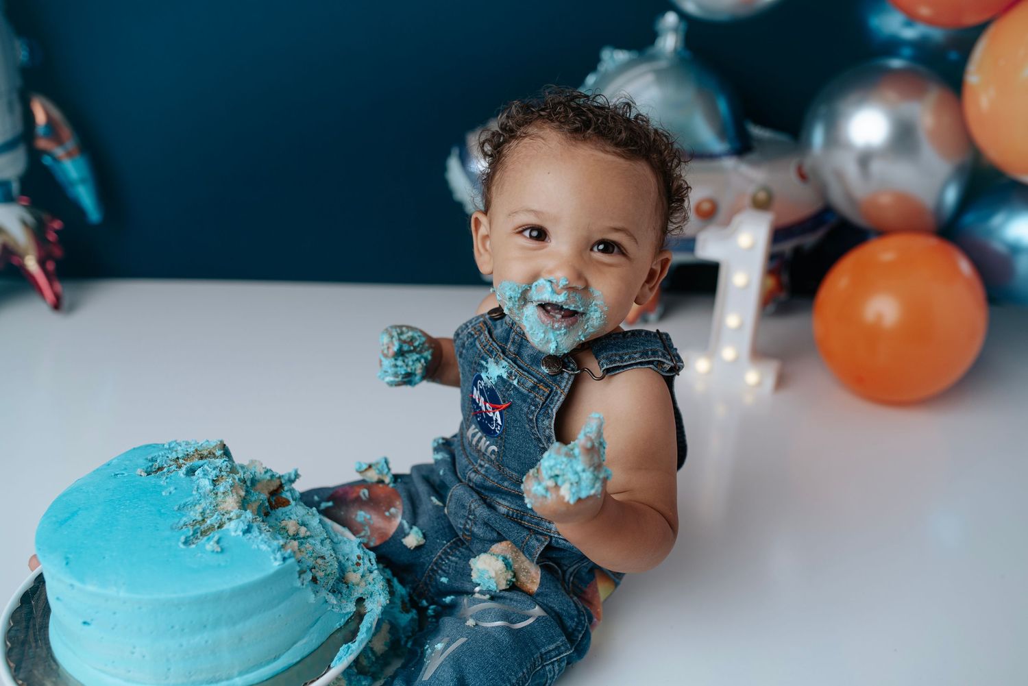 Happy one-year-old boy covered in blue frosting during a cake smash birthday photoshoot with balloons. Ivy Lynn Photography, North Georgia