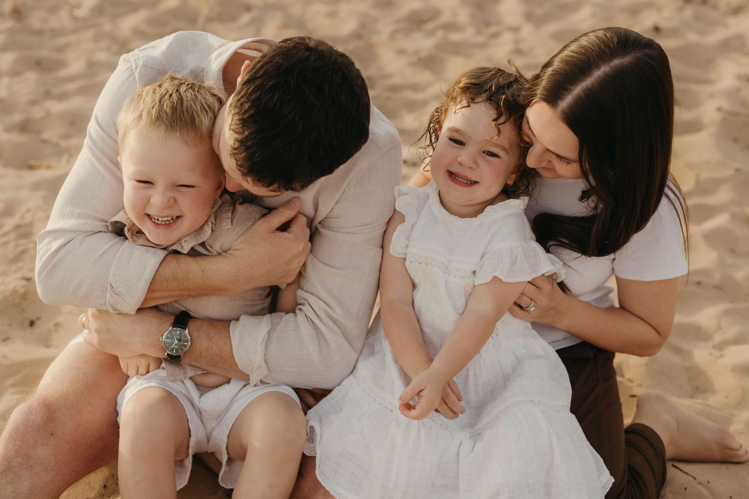 Happy family of four sitting on sandy beach, parents hugging and kissing two young laughing children warmly.