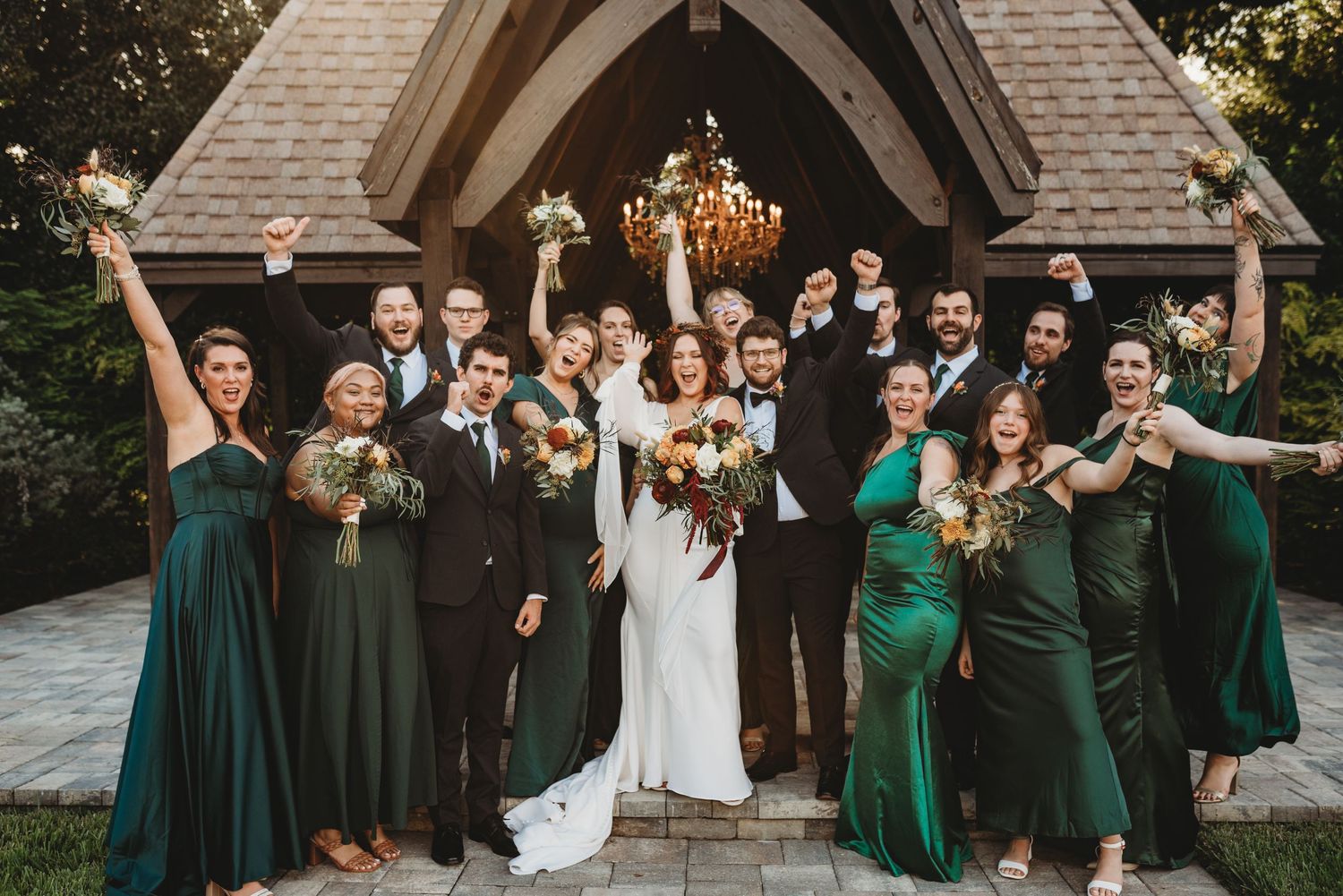 Wedding party celebrating with arms raised outside chapel in emerald green dresses.