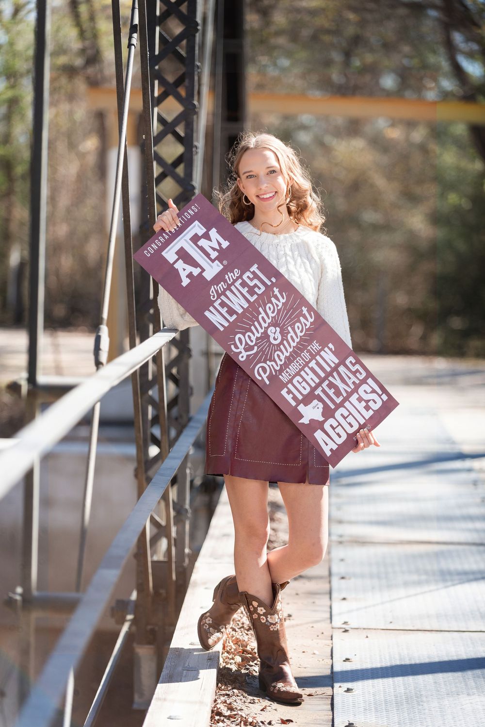 Senior girl sanding on old bridge smiling wearing skirt and boots while holding new Texas A&M acceptance poster