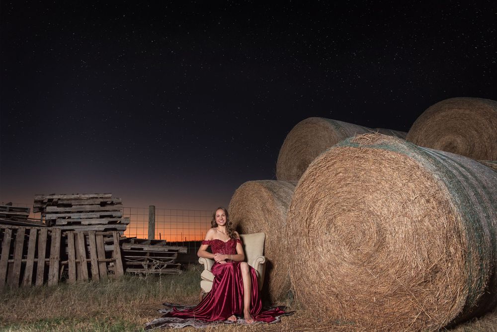 Senior girl wearing prom dress in chair next to haybale at night