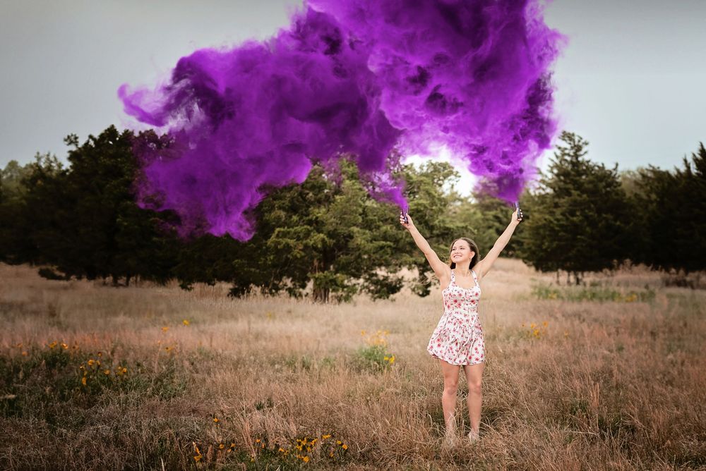 Senior girl in field wearing a floral dress and waving 2 purple smoke bombs