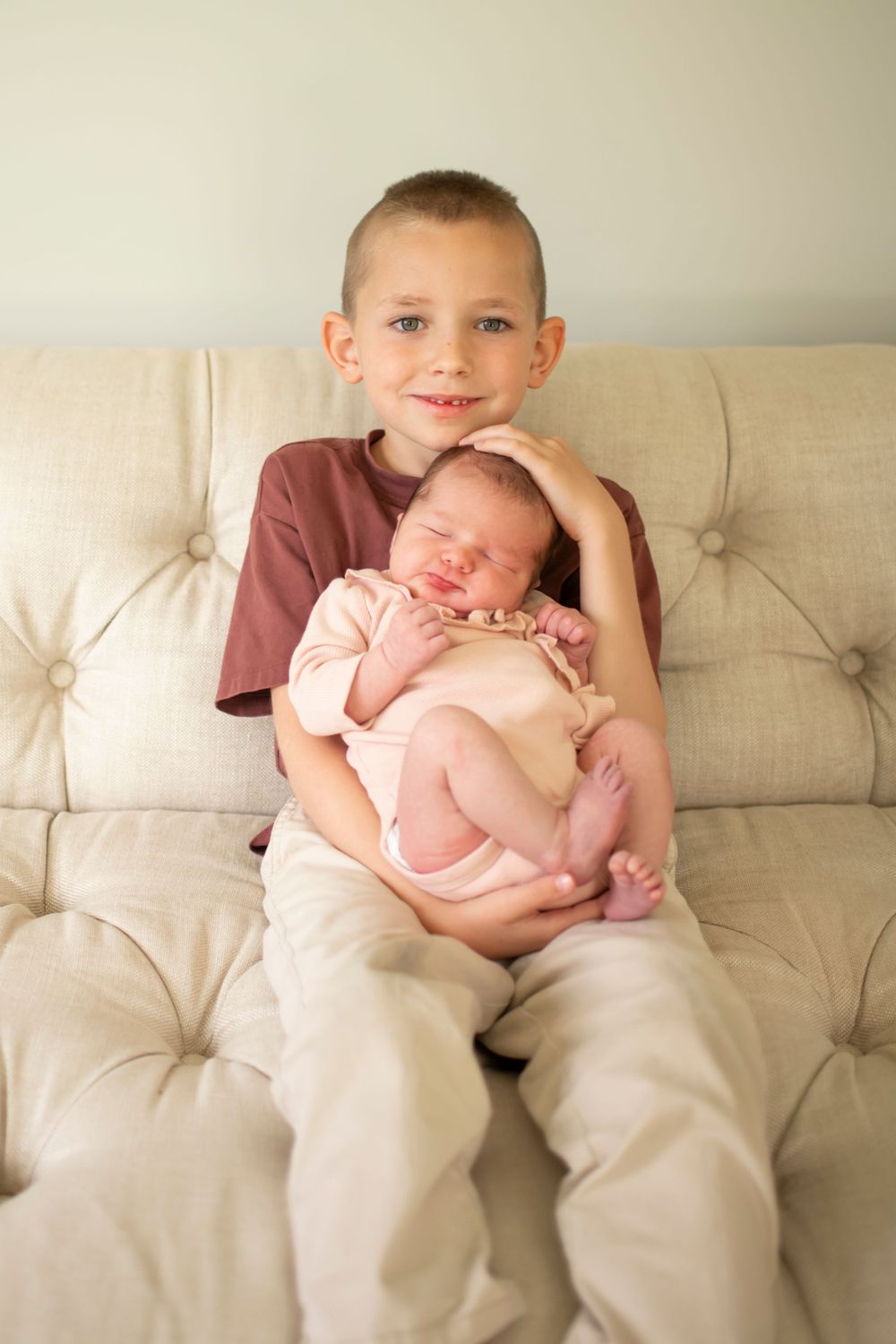 Sweet portrait of siblings sitting together on a beige tufted couch, older child holding newborn baby with care.