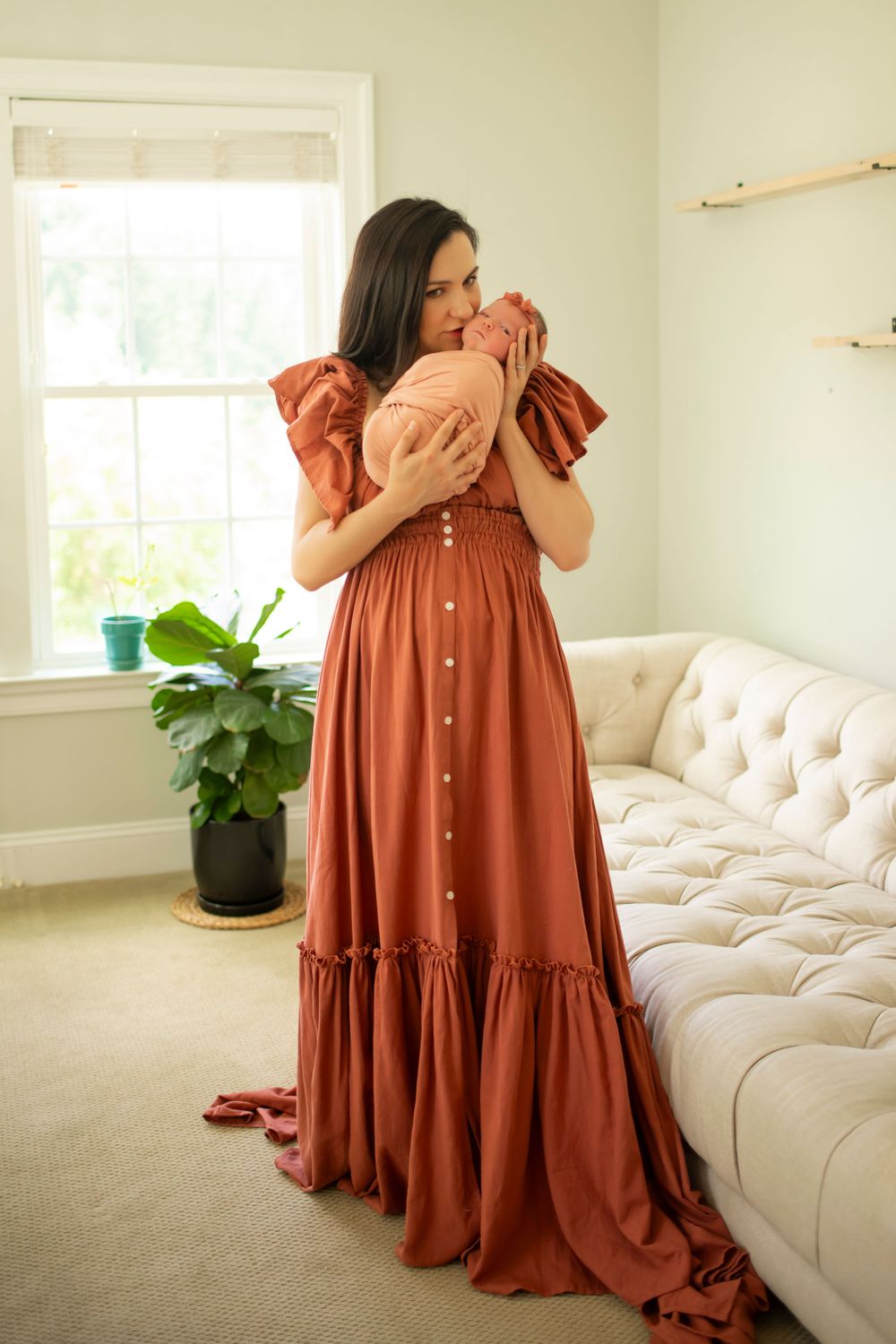 Woman in long rust dress embraces newborn baby near window with houseplant.