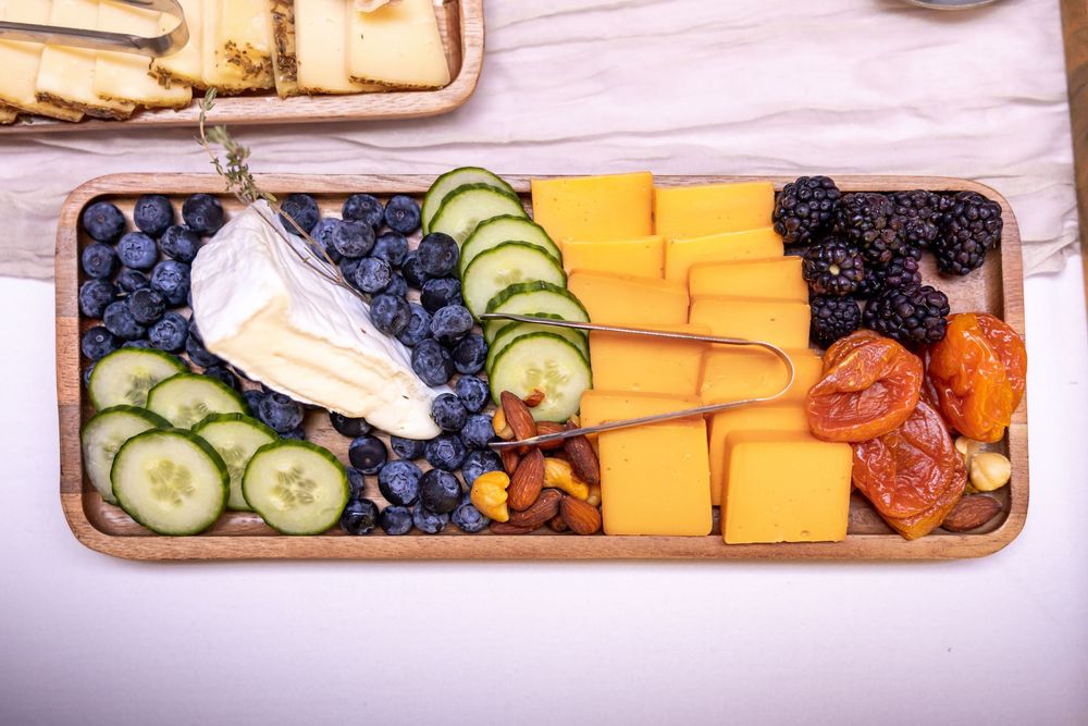 Wooden charcuterie board with cheddar cheese, brie, blueberries, cucumbers, blackberries, dried fruit, and nuts.