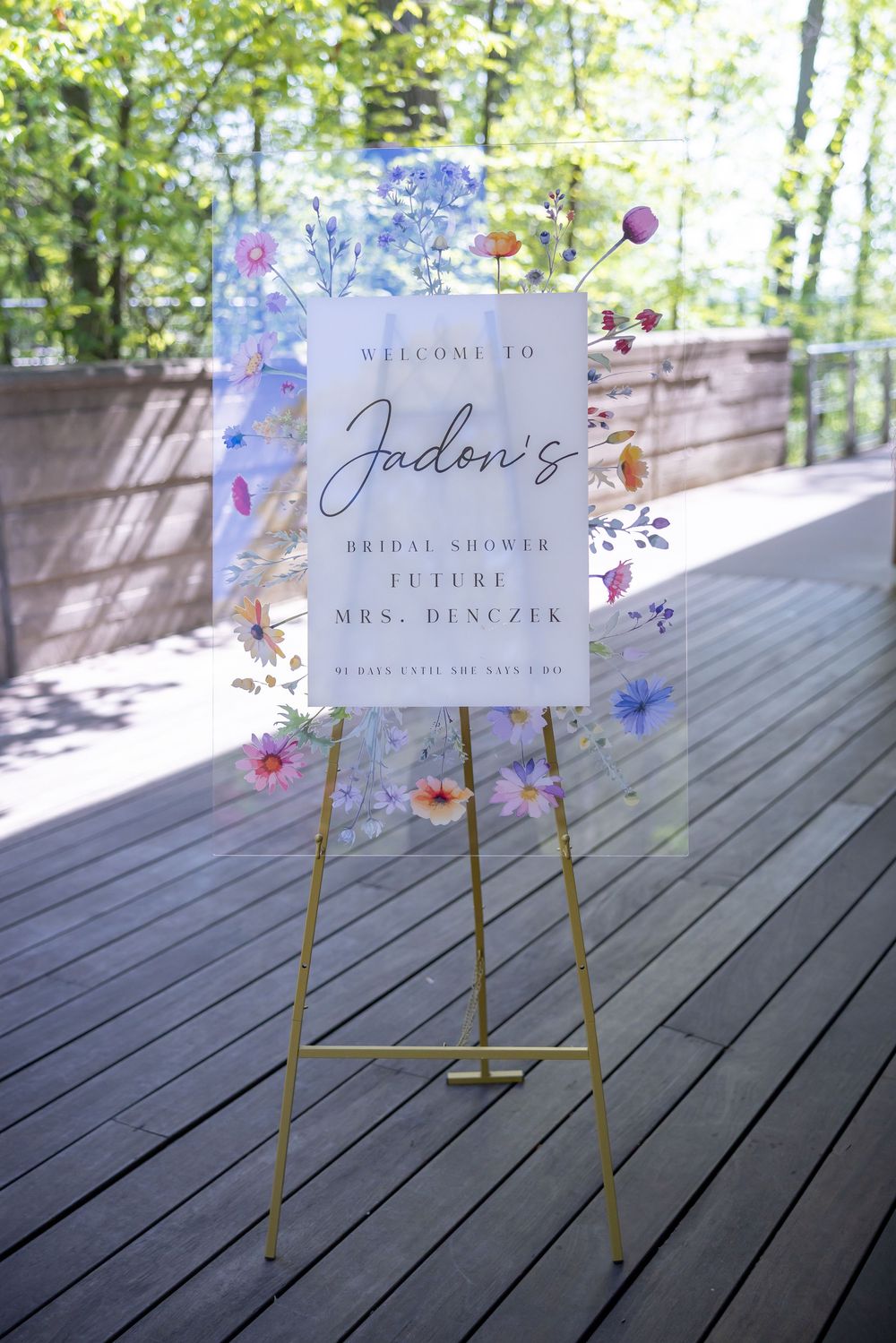 Gold easel with floral acrylic welcome sign for Jadon's bridal shower, future Mrs. Denczek, on a wooden deck.