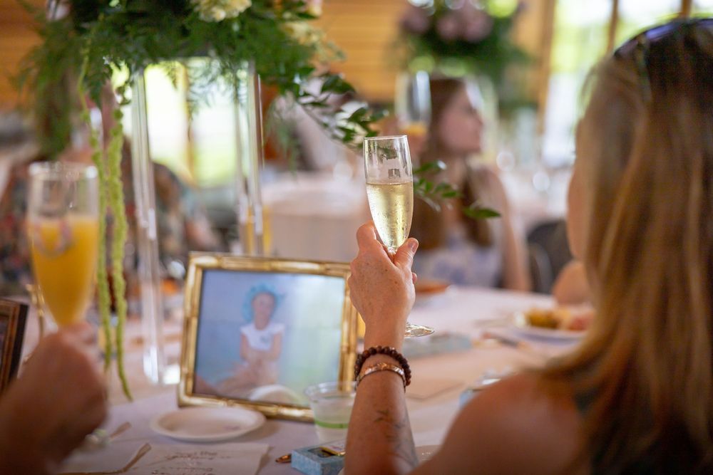 Woman raising a champagne glass in a toast at a wedding reception with floral centerpieces and a framed photo on the table.