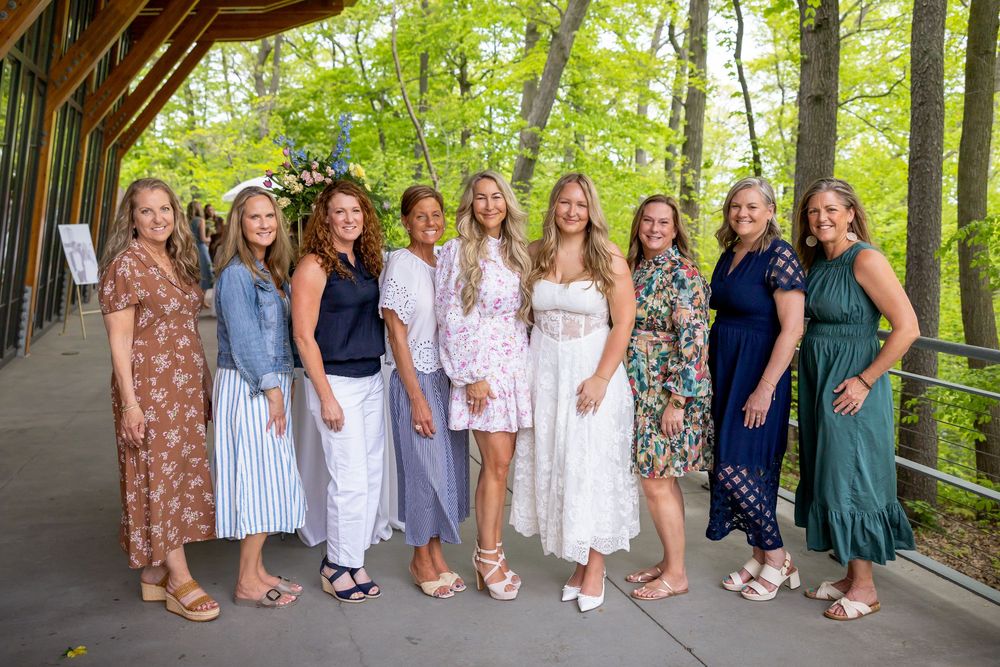 Group of nine women in summer dresses smiling together at outdoor bridal brunch in wooded venue.