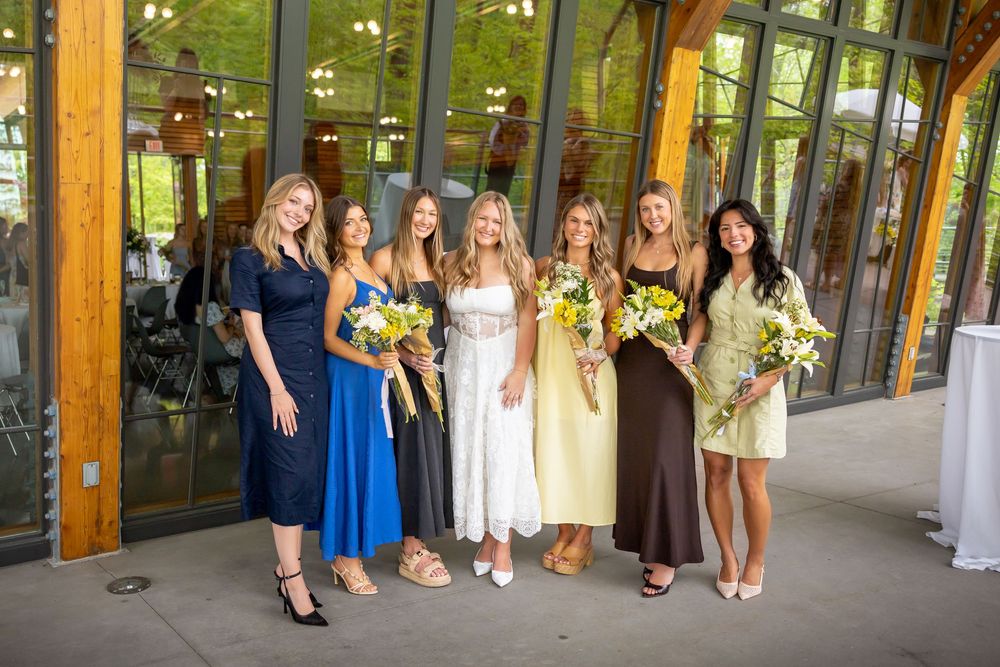 Six women in colorful dresses pose together holding yellow floral bouquets at an elegant indoor wedding venue.