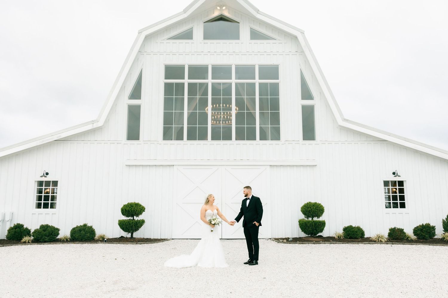 Modern white barn-style event venue with farmhouse architecture features wraparound porch and dormer windows on a grassy field.